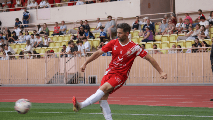 A Barbagiuans Monaco player wearing a red kit featuring the Moravia Yachting logo takes a shot during the Racing Stars Football Cup 2026 at Stade Louis-II, Monaco, with a crowd of spectators in the stands behind him.