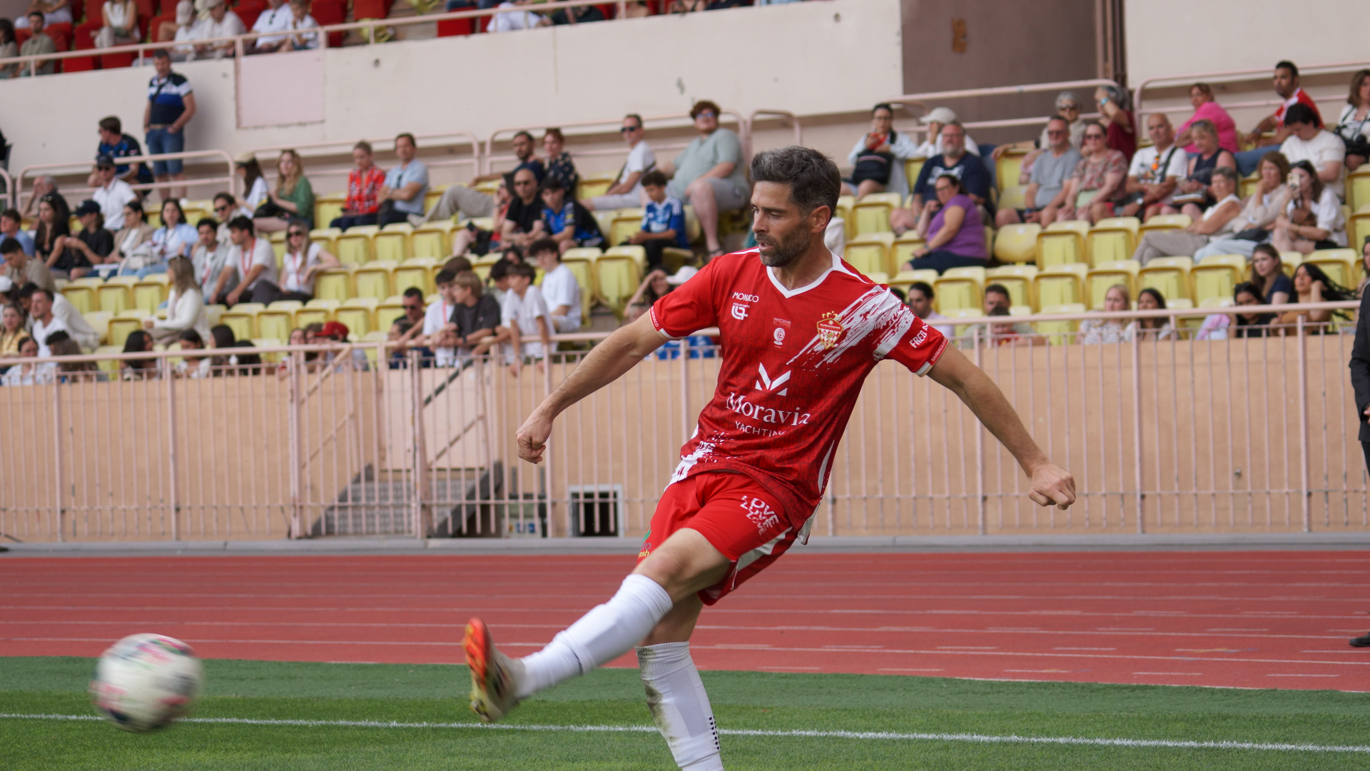 A Barbagiuans Monaco player wearing a red kit featuring the Moravia Yachting logo takes a shot during the Racing Stars Football Cup 2026 at Stade Louis-II, Monaco, with a crowd of spectators in the stands behind him.